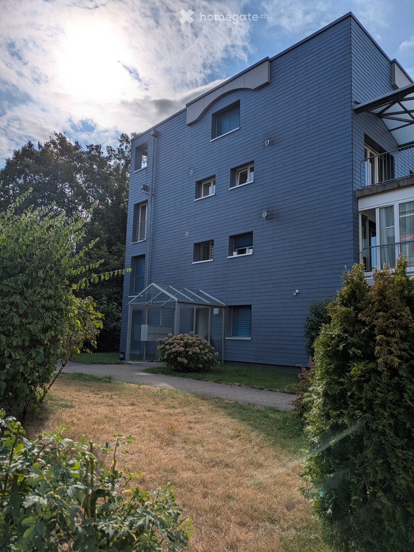 Modern two-story house with grey siding, multiple windows, and a covered entrance area. The house features a well-maintained garden with shrubs and trees.