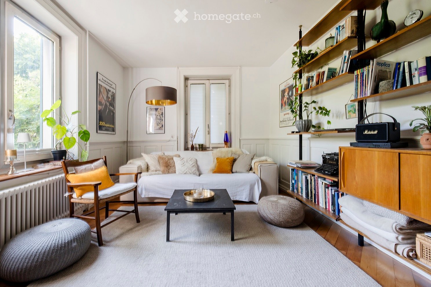 A cozy living room with a white sofa, coffee table, wooden chair, floor lamp, and shelves filled with books and plants. The room features large windows with a view of greenery outside.
