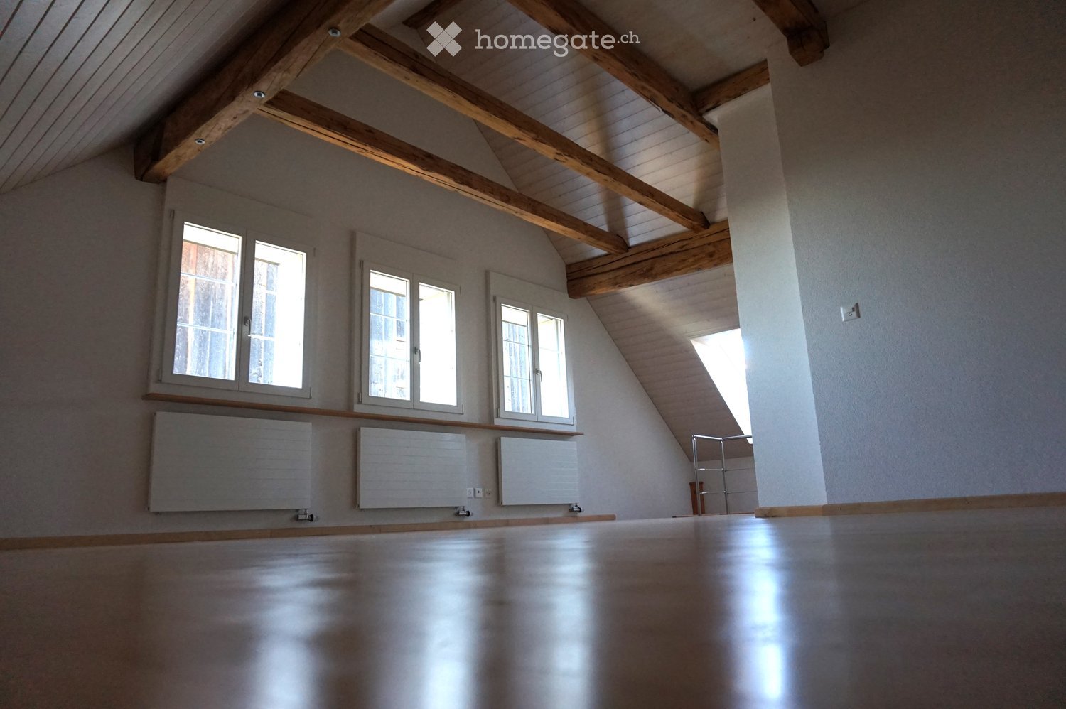 An empty attic room with wooden beams, exposed wooden ceiling, large windows, and radiators. The floor is polished wood, and the walls are painted white.