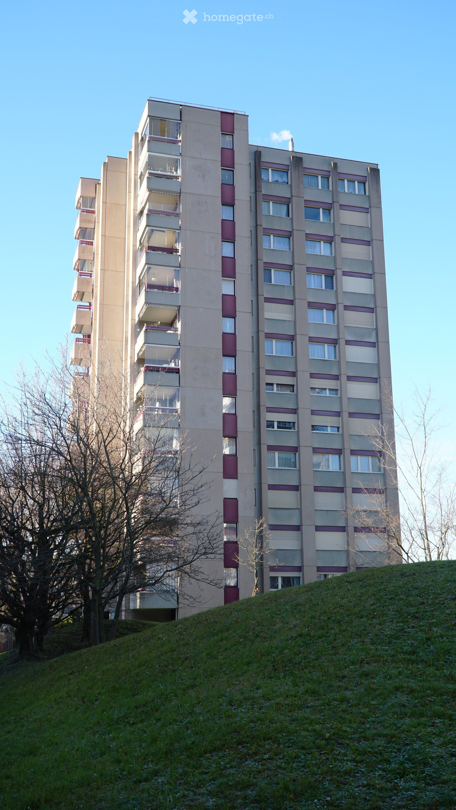 Multi-story residential building with balconies and windows, situated on a grassy slope with trees nearby.