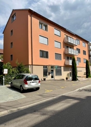 Four-story apartment building with orange facade, multiple windows, and balconies. A silver car is parked in front of the building.