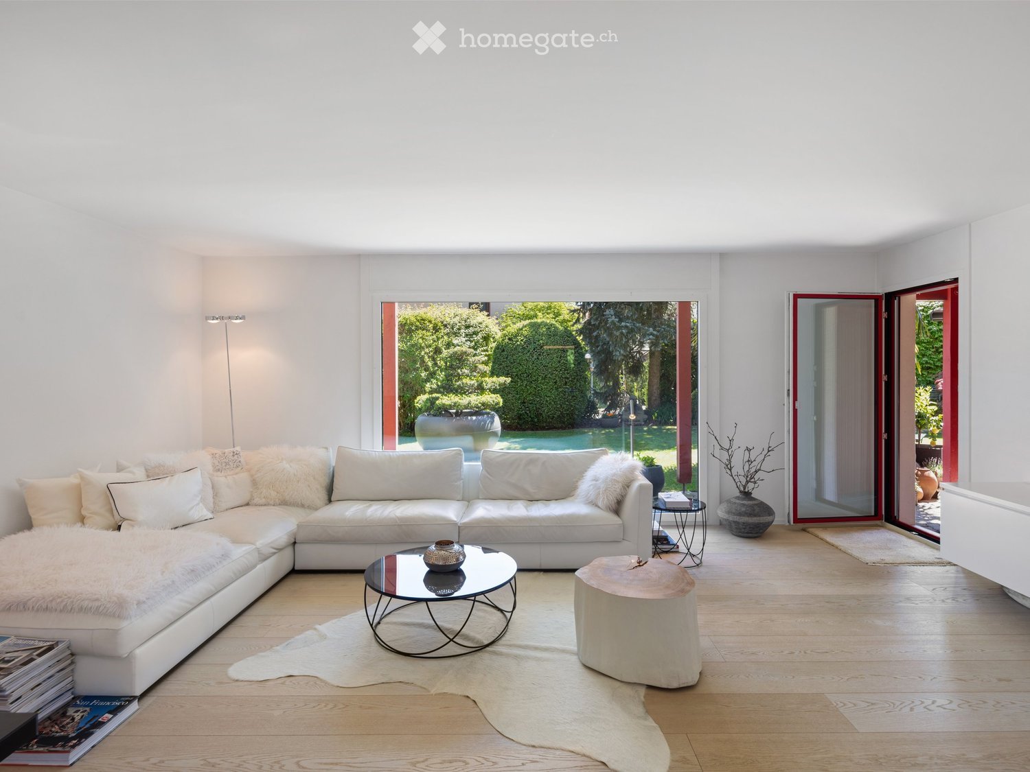 Modern living room with white sectional sofa, glass coffee table, wooden floor, and large windows opening to a garden.