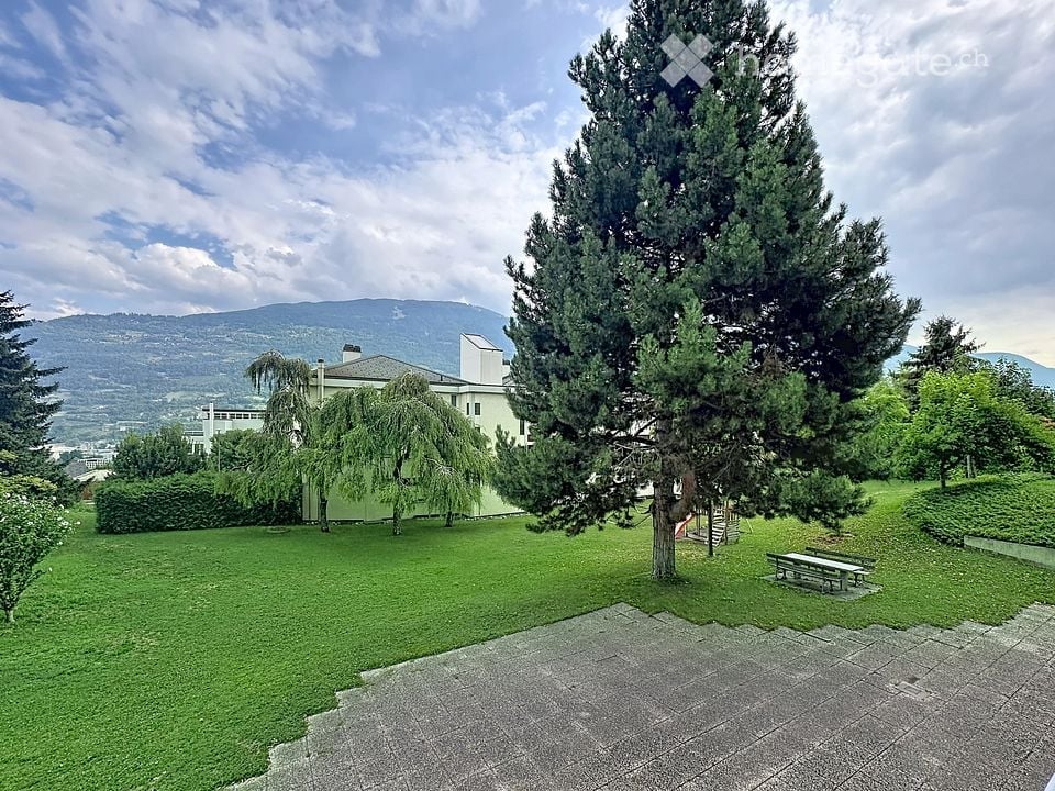 Well-maintained garden with green lawn, tall trees, and a view of mountains in the background.