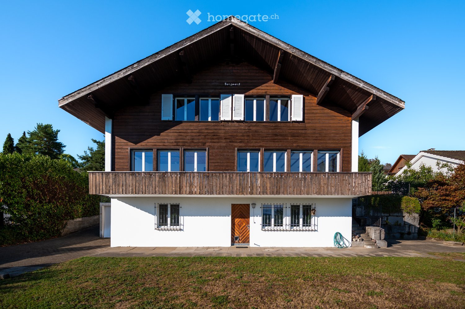 Two-story wooden house with a steeply pitched roof, large windows, and a balcony on the upper level. The lower level has a white facade with a central wooden door and metal-framed windows.