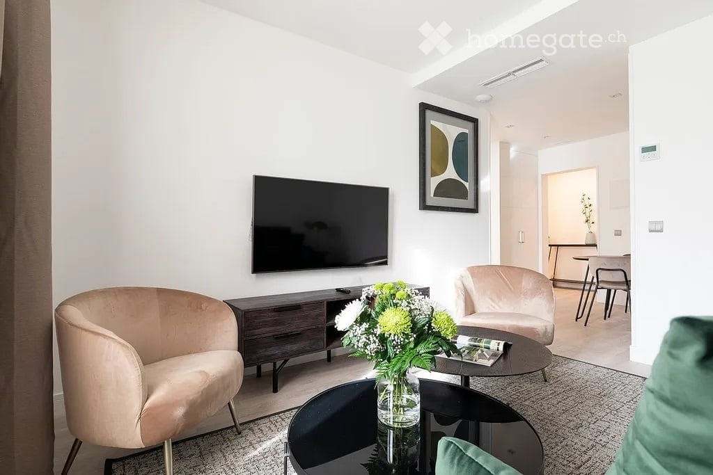 Modern living room with a sleek black TV mounted on the wall, a dark wooden TV stand, a round coffee table with a flower arrangement, two light-colored armchairs, and a dining area visible in the background.