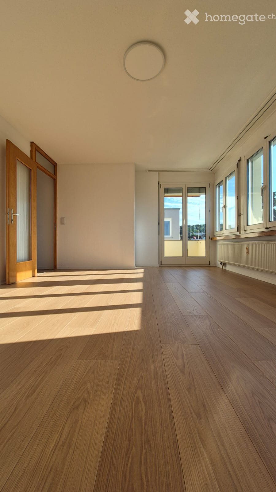 Empty room with wooden flooring, large windows, and a sliding glass door leading to a balcony.