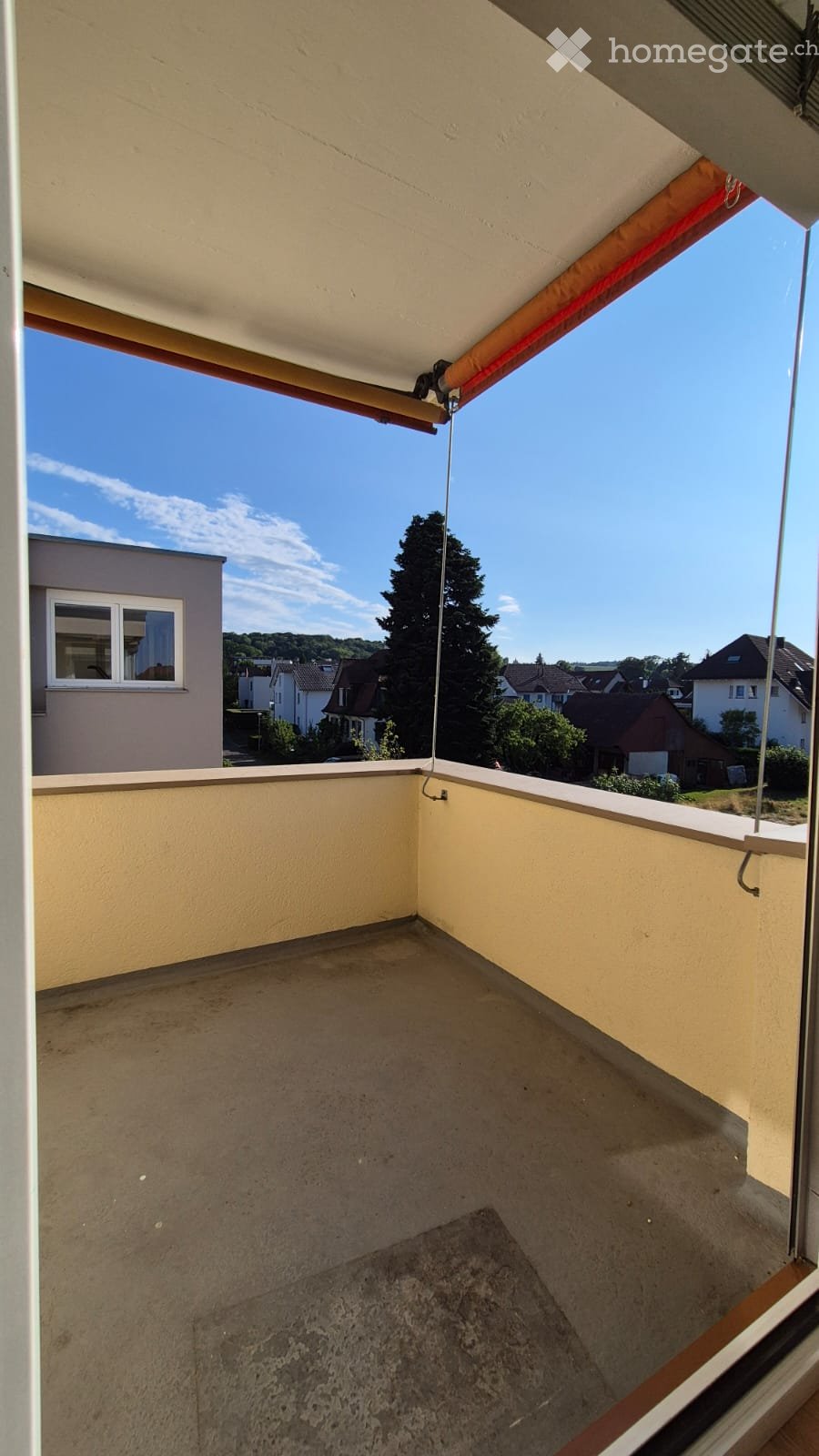 Empty balcony with concrete floor, beige walls, and a retractable awning. Offers a view of neighboring houses and trees.