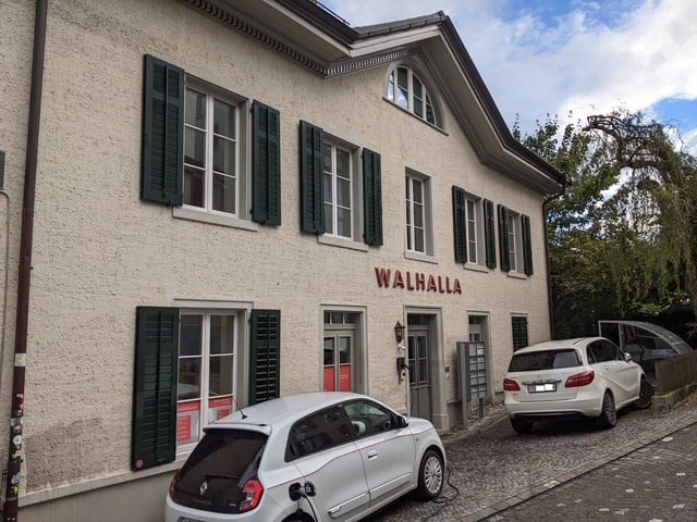 A two-story building with a light-colored facade, green shutters, and the name "WALHALLA" displayed on the front. Two cars are parked in front of the building.