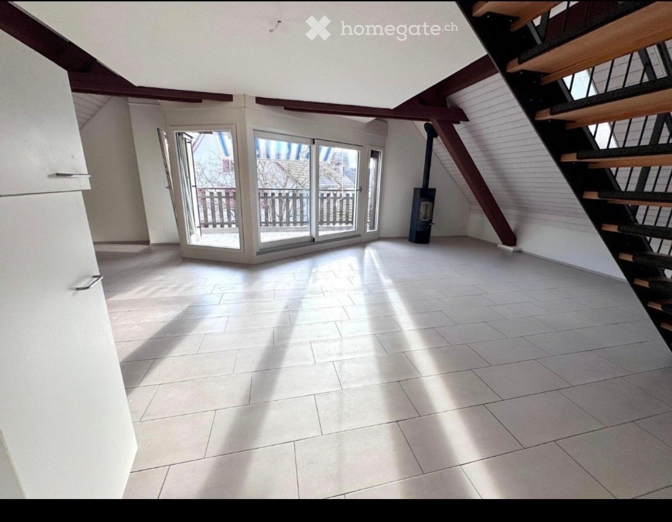 Empty room with white tiled floor, exposed wooden beams, staircase leading upwards, and large glass doors opening to a balcony area.
