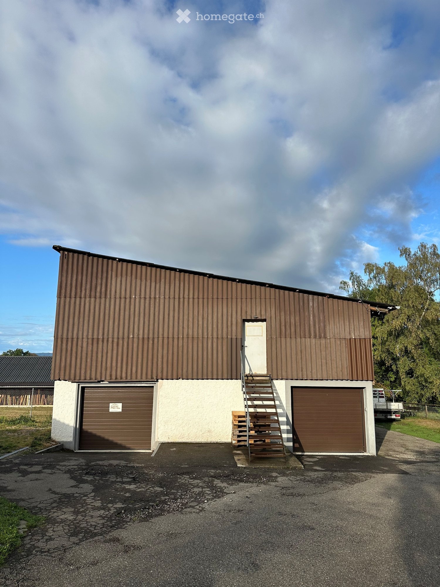 A single-story industrial-style building with a brown metal roof and white lower walls, featuring two large garage doors and a small staircase leading to an upper-level entrance.