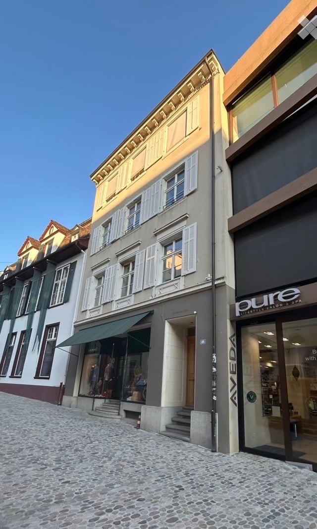 A multi-story building with classic European architecture, featuring a mix of beige and grey facades, white shutters, and a green awning over the entrance. The building is part of a row of similar structures on a cobblestone street.
