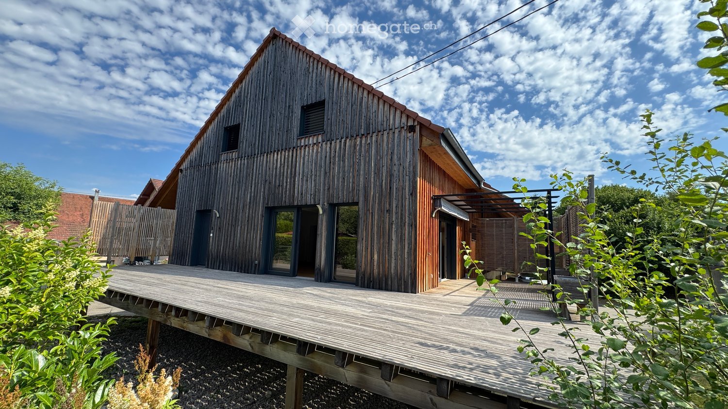 Modern wooden house with a raised ground floor, large windows, and a wooden deck extending from the front entrance.
