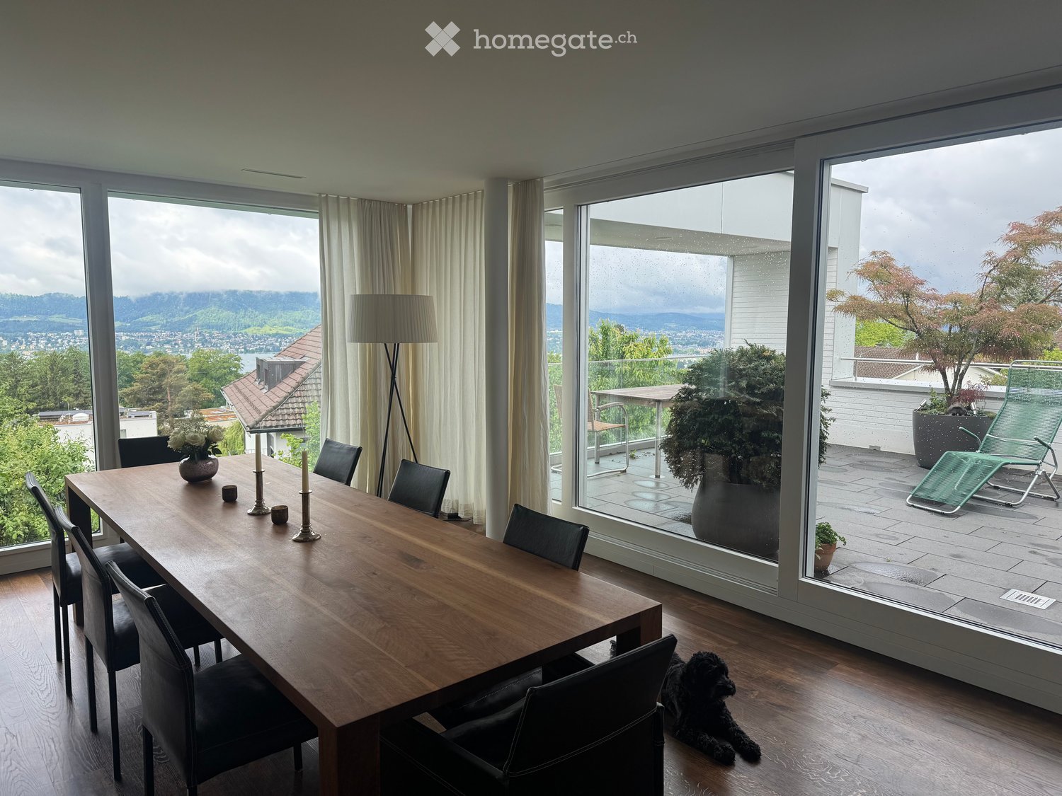 Dining room with a large wooden table, black chairs, a vase with white flowers, and a floor lamp. Large windows offer a scenic view of the landscape, with a small balcony area visible through the glass.