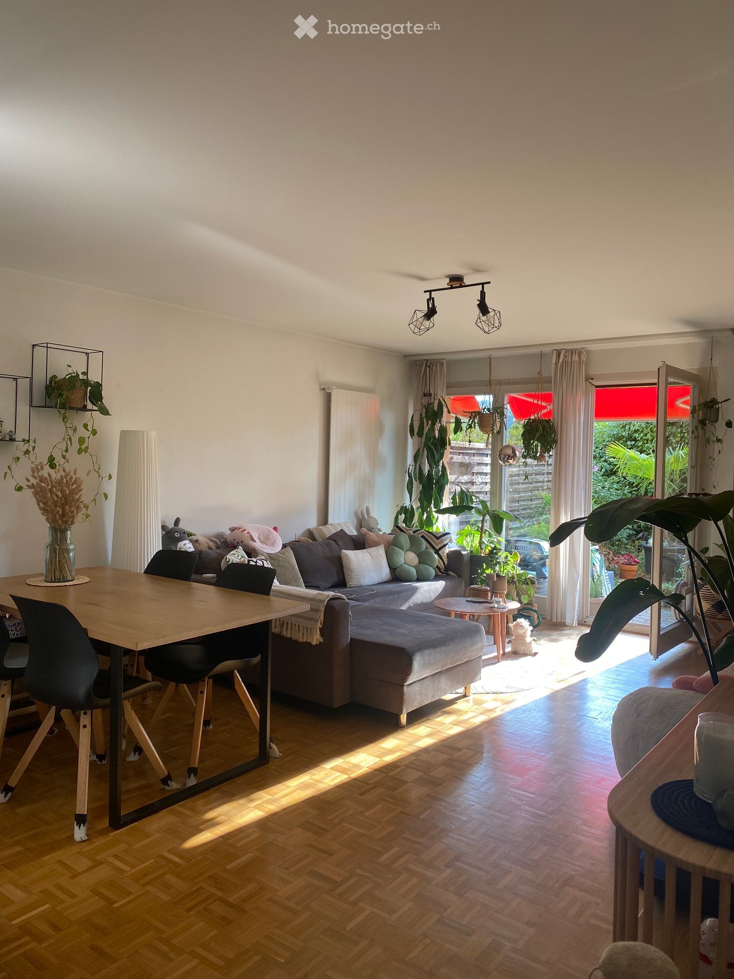Bright living room with wooden flooring, large window with sheer curtains, and a sliding glass door leading to a garden. Features a grey sectional sofa with multiple cushions, a wooden coffee table, a dining table with chairs, and several potted plants.