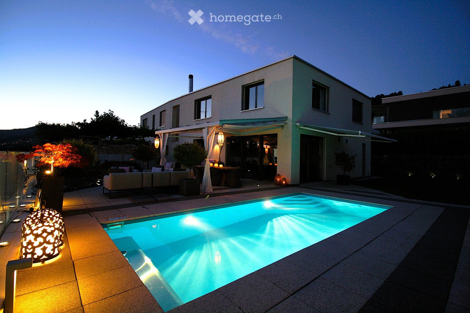 Modern outdoor area with a swimming pool, patio furniture, and ambient lighting, featuring a contemporary house in the background.