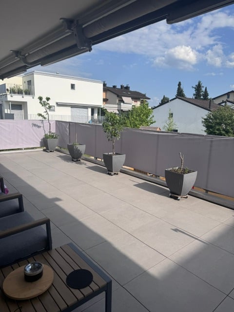 Modern balcony with grey tiles, grey furniture including a small table and chairs, several potted plants, and a grey privacy fence.