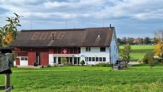 A large, traditional Swiss farmhouse with a red barn and white main building, surrounded by a green field and trees.
