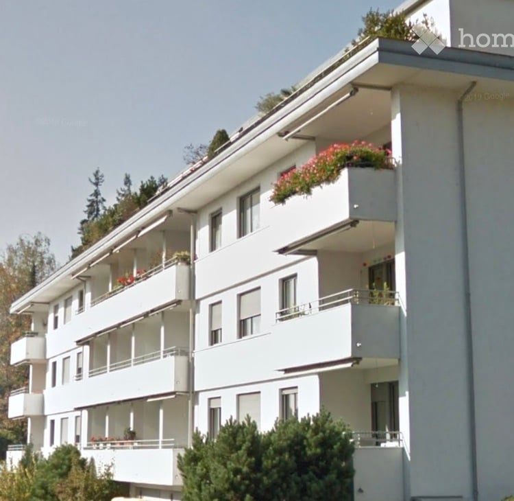 Modern multi-story apartment building with white balconies and flower boxes, surrounded by greenery.