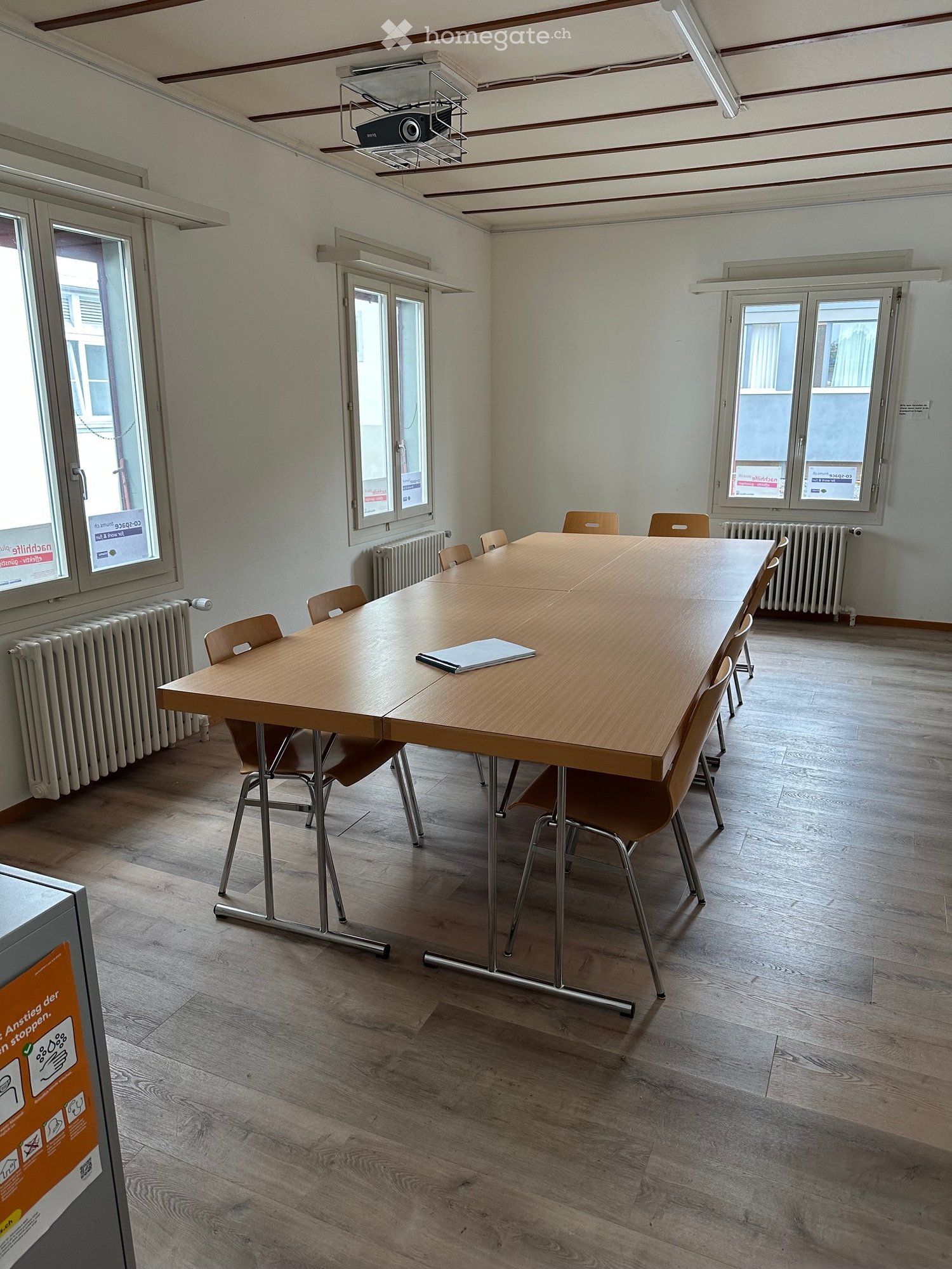Meeting room with a long wooden table, chairs, radiator, and a projector mounted on the ceiling.