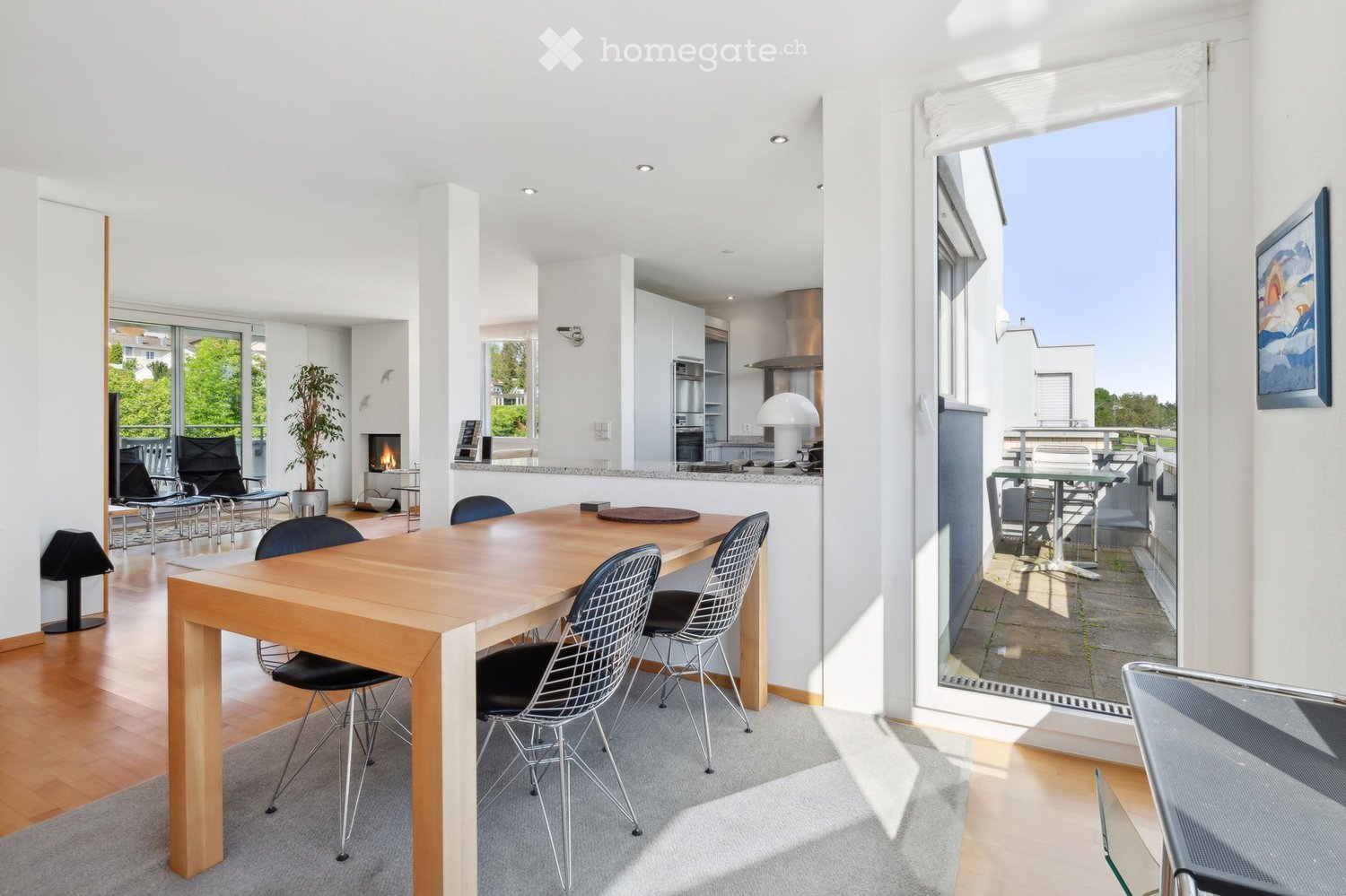 Open-plan living and dining area with a wooden dining table, black wire chairs, a fireplace, a modern kitchen with stainless steel appliances, and a balcony with outdoor furniture.
