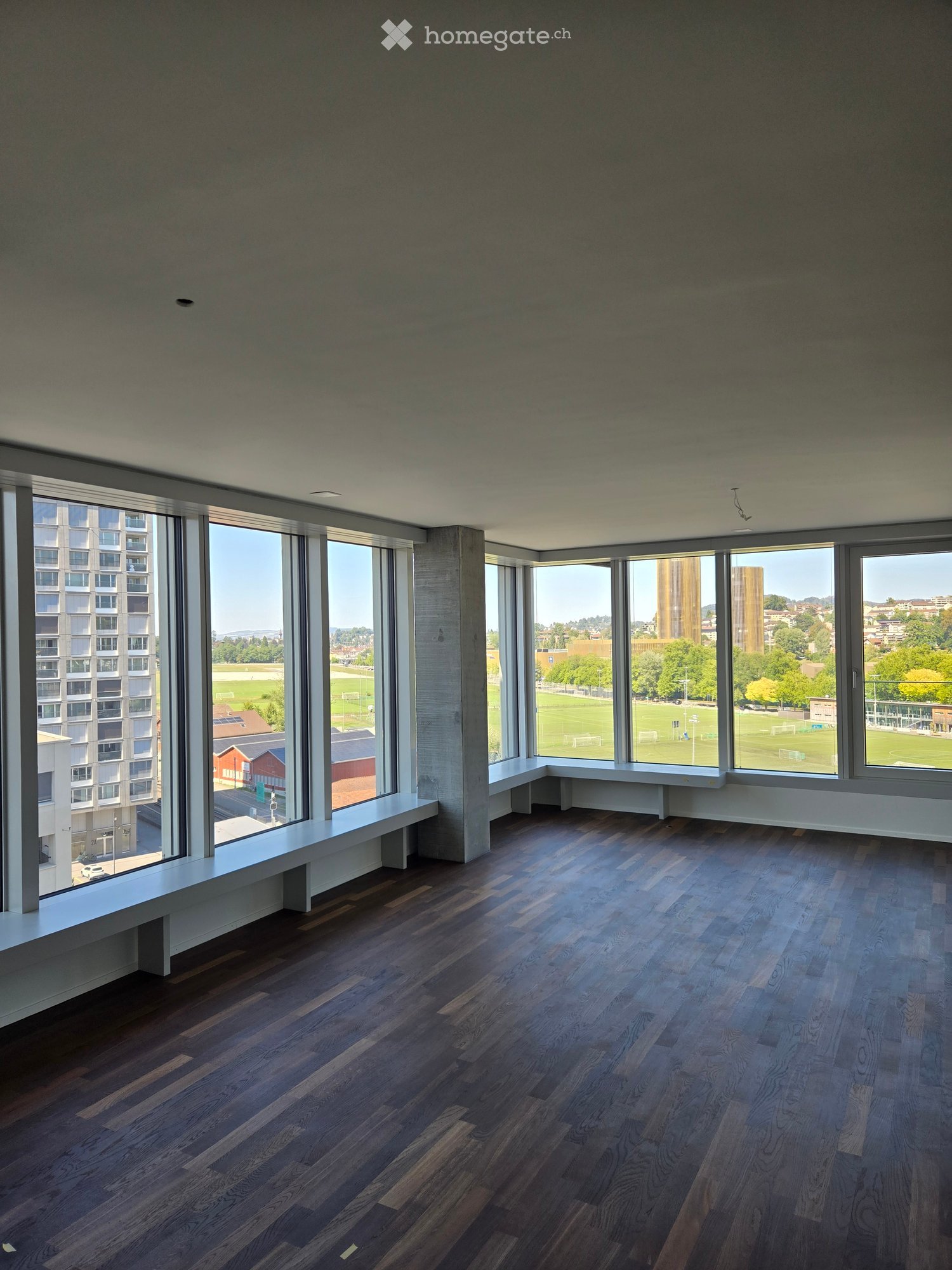 Empty room with dark wooden flooring, large windows offering a view of a park and buildings, and built-in benches along the windows.