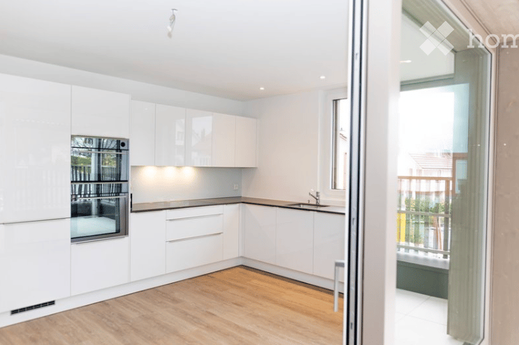 Modern, minimalist kitchen with white cabinetry, a built-in oven, and a sleek sink. The space features light wood flooring and an open sliding door leading to a balcony.