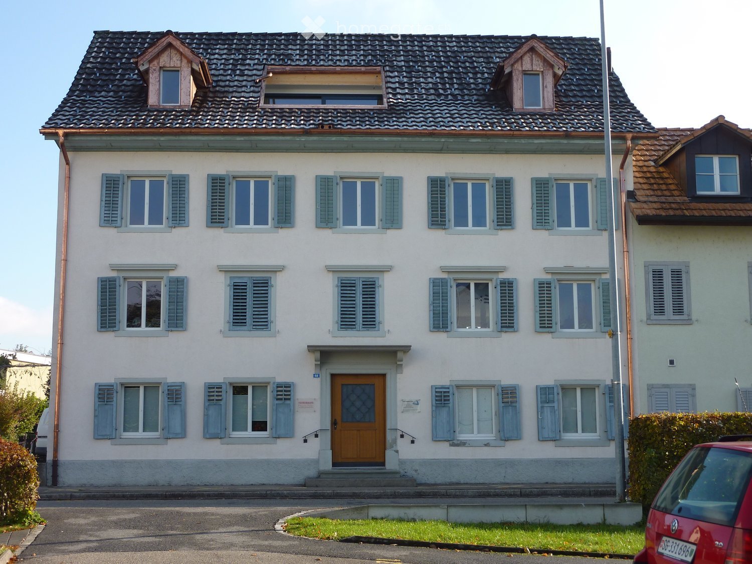 Two-story house with light-colored walls, multiple windows with shutters, and a central wooden front door.