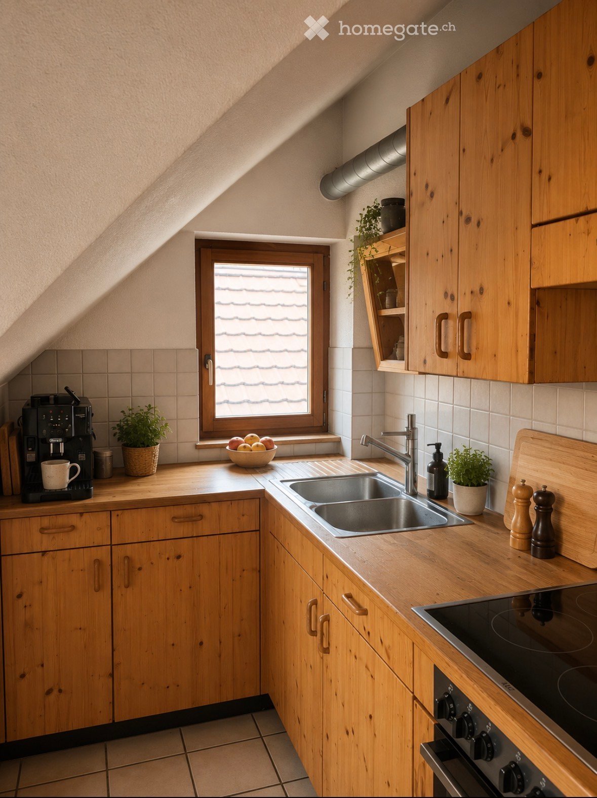 A cozy attic kitchen with wooden cabinets, a stainless steel sink, and a modern electric stove. The space features a window with a view of the roof, a coffee maker, and small potted plants on the countertops.