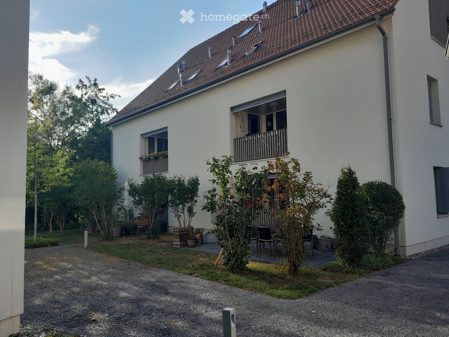 Two-story house with white exterior, brown tiled roof, and balcony on the upper floor. The garden area includes several potted plants, a small seating area with chairs, and a gravel pathway.