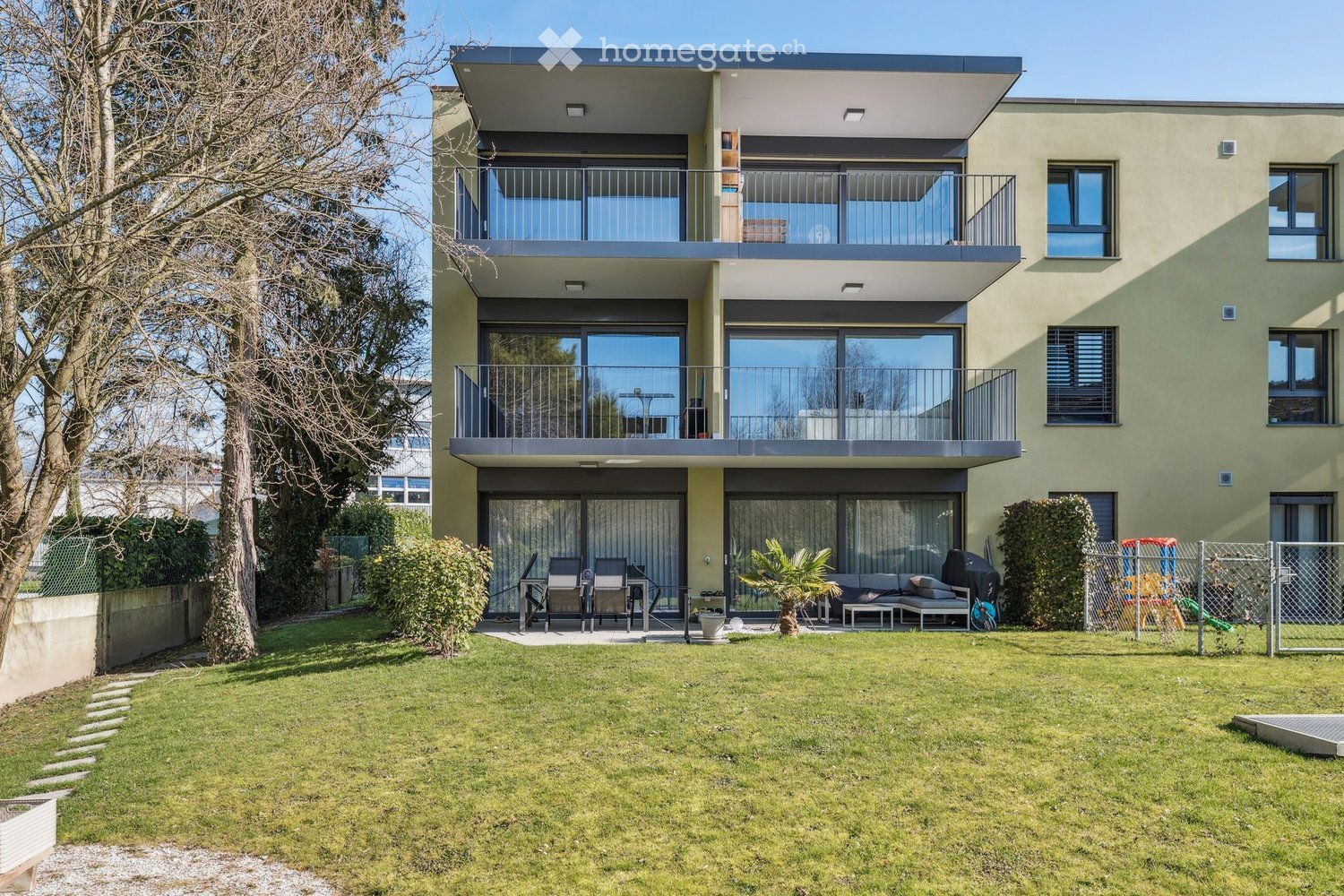Modern two-story house with large windows and balconies, surrounded by a well-maintained lawn and outdoor seating area.