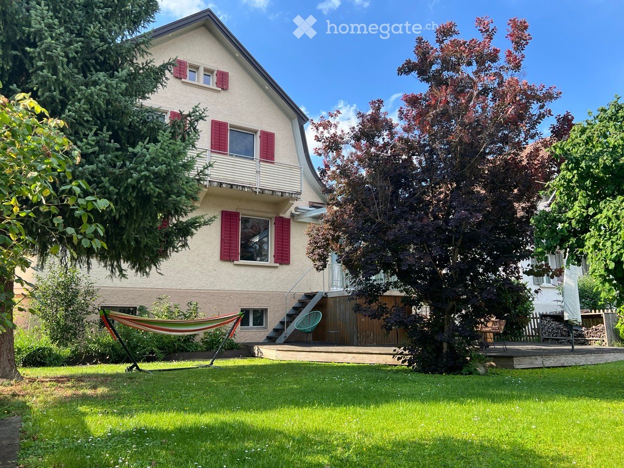 Two-story house with red shutters, white balcony railings, and a well-maintained garden with a hammock, patio, and trees.