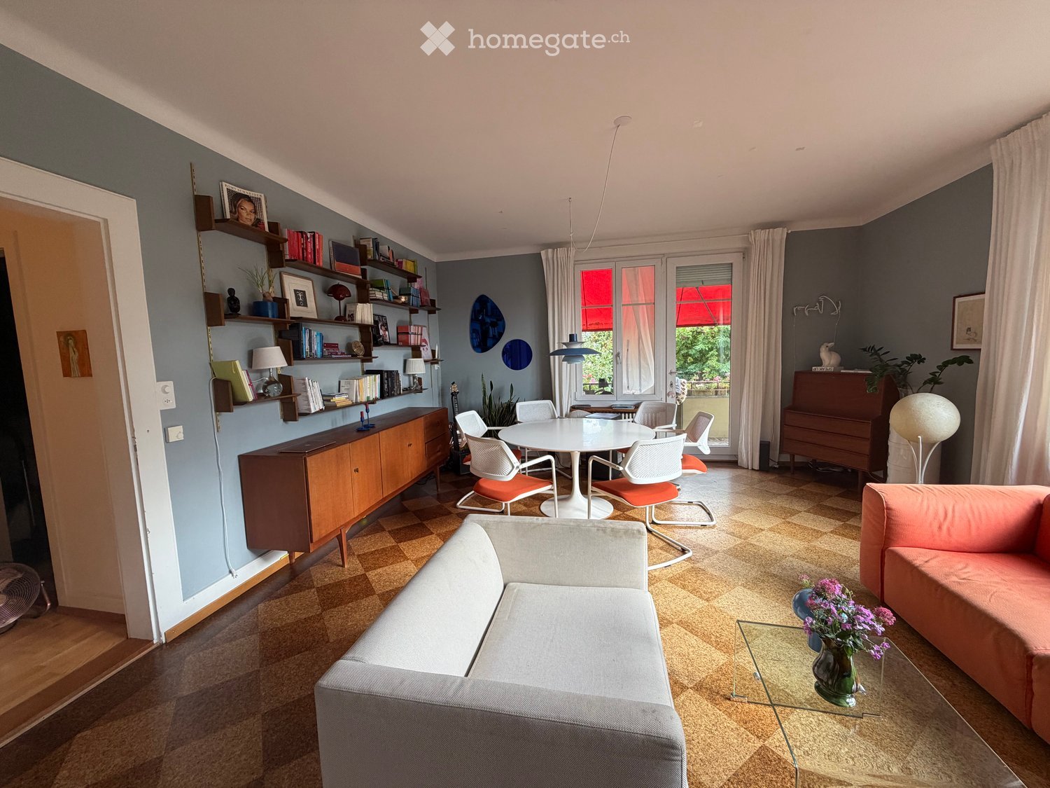 A modern living room with a grey sofa, orange armchair, white dining table with chairs, wooden cabinet, shelves with books and decor, and a piano. The room features a checkered floor and large windows with curtains.