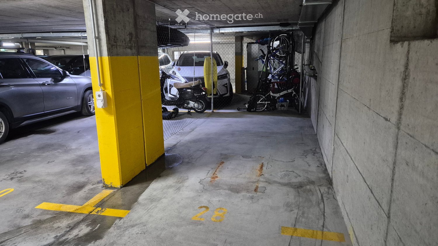 An indoor parking garage with a numbered parking space, a parked car, a parked motorcycle, a rack with bicycles, and a power outlet on the wall.