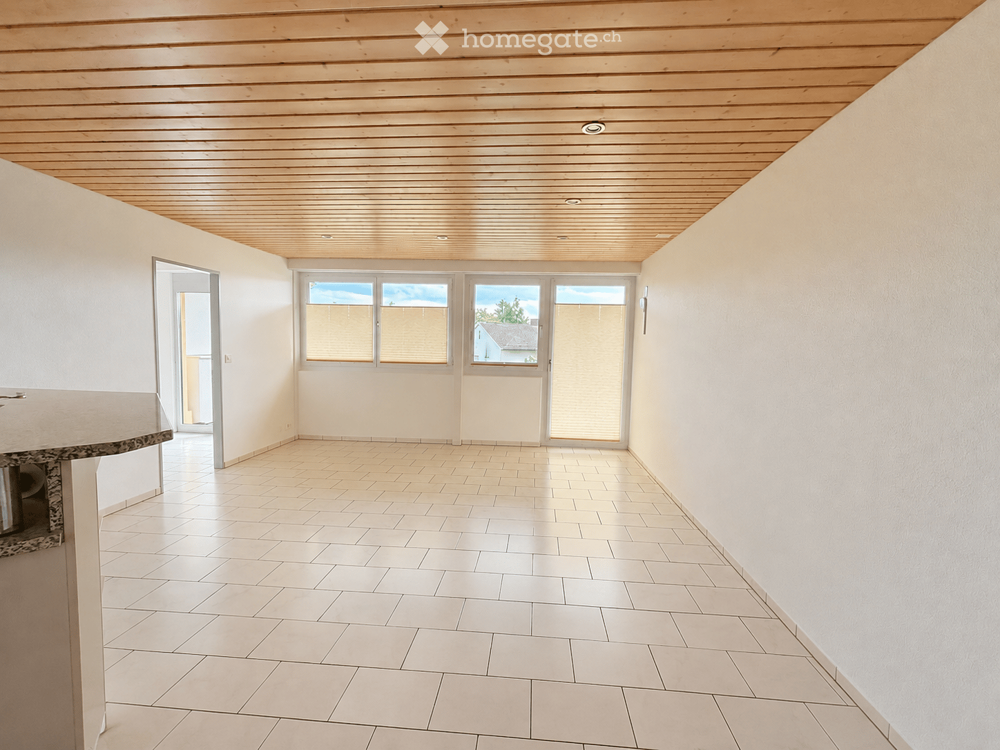 Empty room with white tiled floor, light-colored walls, and a wooden slatted ceiling. Features large windows with blinds and a doorway leading to another space.