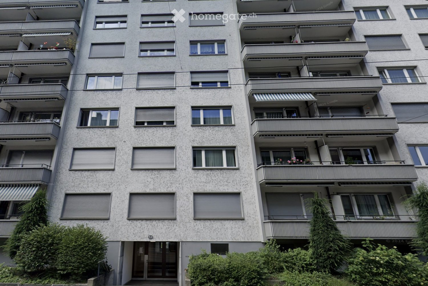 Multi-story apartment building with multiple balconies, large windows, and greenery around the base.