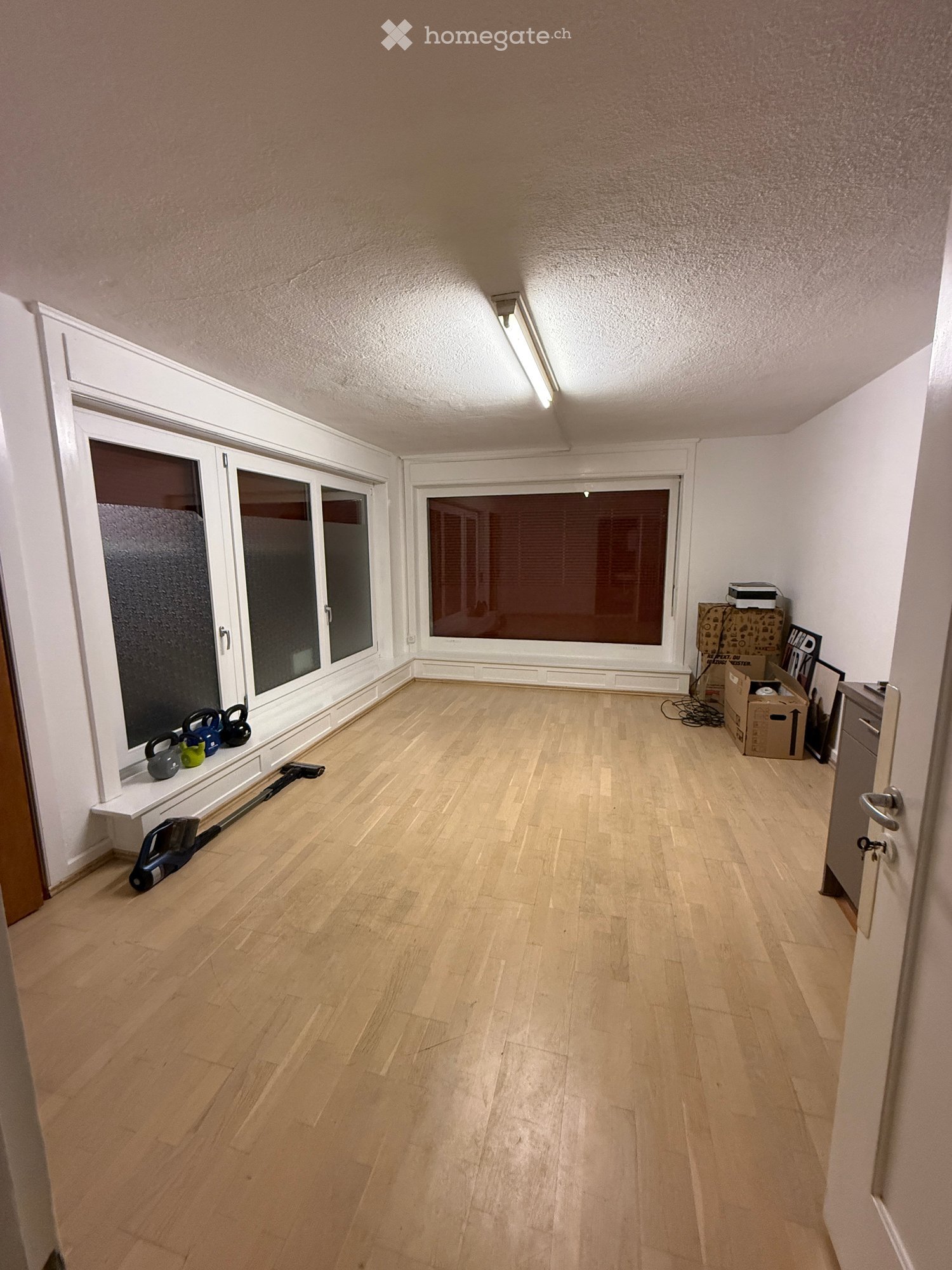 Empty room with wooden flooring, white walls, large windows, and a small shelf with a few items on it.
