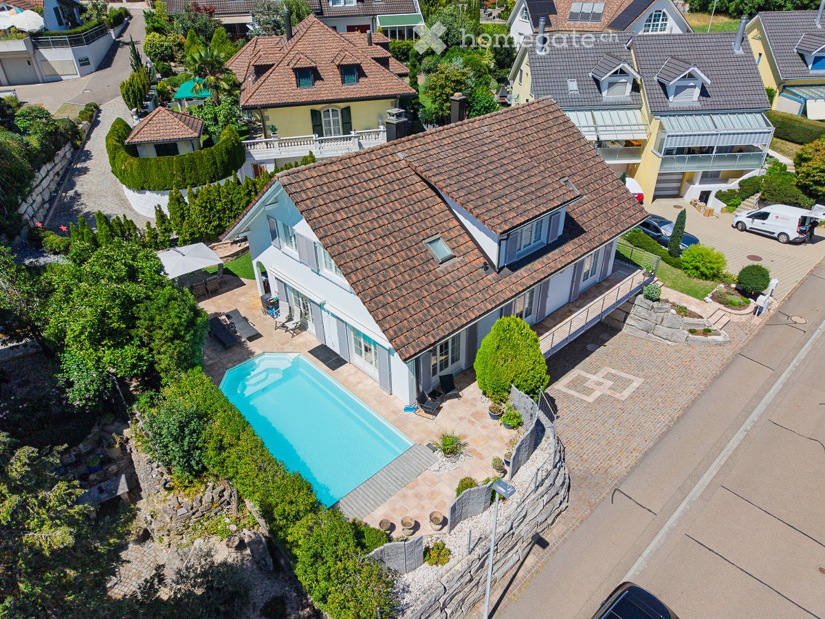 Aerial view of a two-story house with a tiled roof, surrounded by greenery and a swimming pool in the garden area.