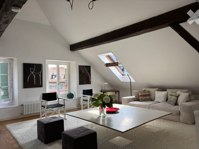 Bright living room with slanted wooden ceiling beams, large windows, and skylight. Features a grey sofa, coffee table, two black chairs, two dark grey ottomans, a small side table with a lamp, and decorative art pieces on the walls.