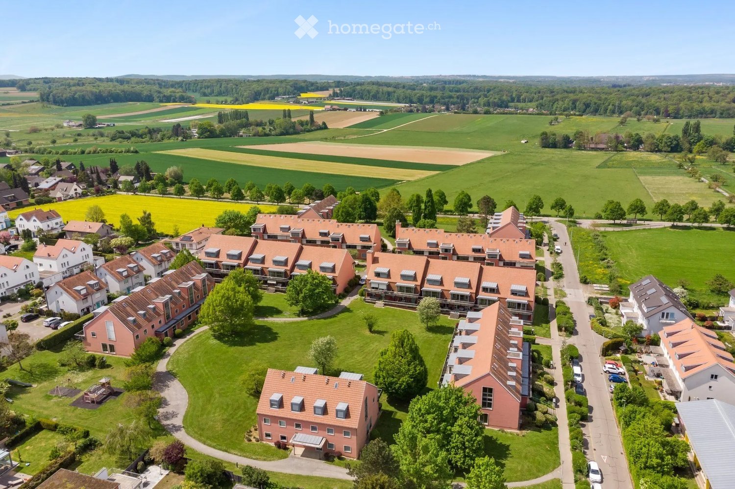 Aerial view of a residential area with multiple houses, green lawns, trees, and surrounding farmland.