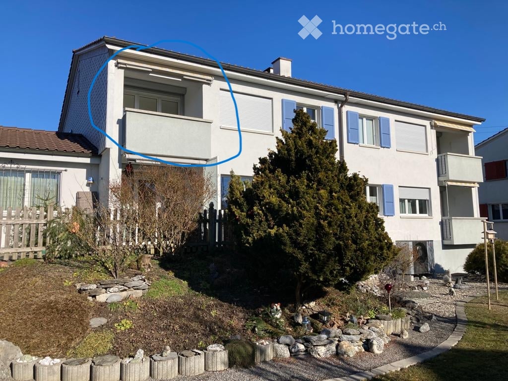 Two-story house with white exterior, blue shutters, and balconies on both floors. The front yard includes a small garden with rocks and plants.