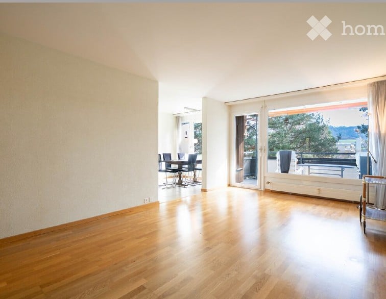 Bright, open living space with wooden flooring, large sliding glass doors leading to a balcony, and a small dining area with a table and chairs.