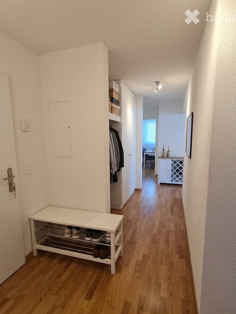 A well-lit hallway with wooden flooring, white walls, and a built-in shoe rack. A small bench with shoes is visible, and a closet with hanging clothes is on the right side. The hallway leads to another room with a dining area visible in the background.