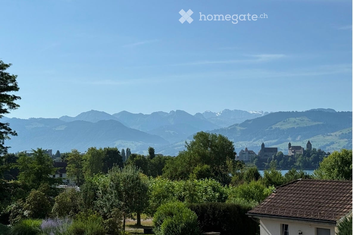 Scenic view of a lake and distant mountains, surrounded by trees and a small house in the foreground.