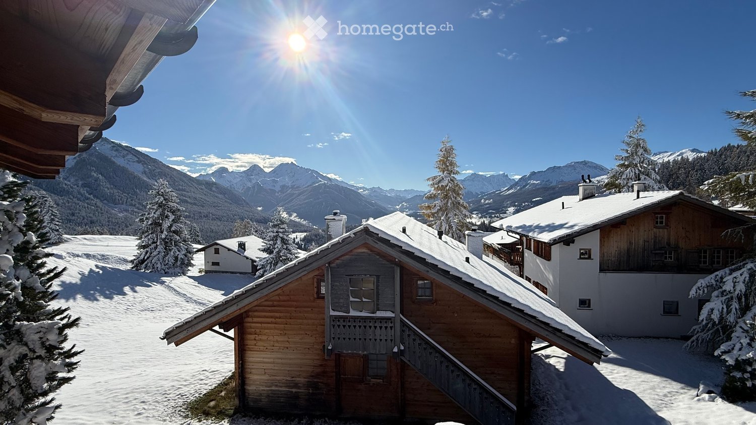 A wooden chalet with snow-covered roof and surrounding snow-covered trees and mountains in the background.