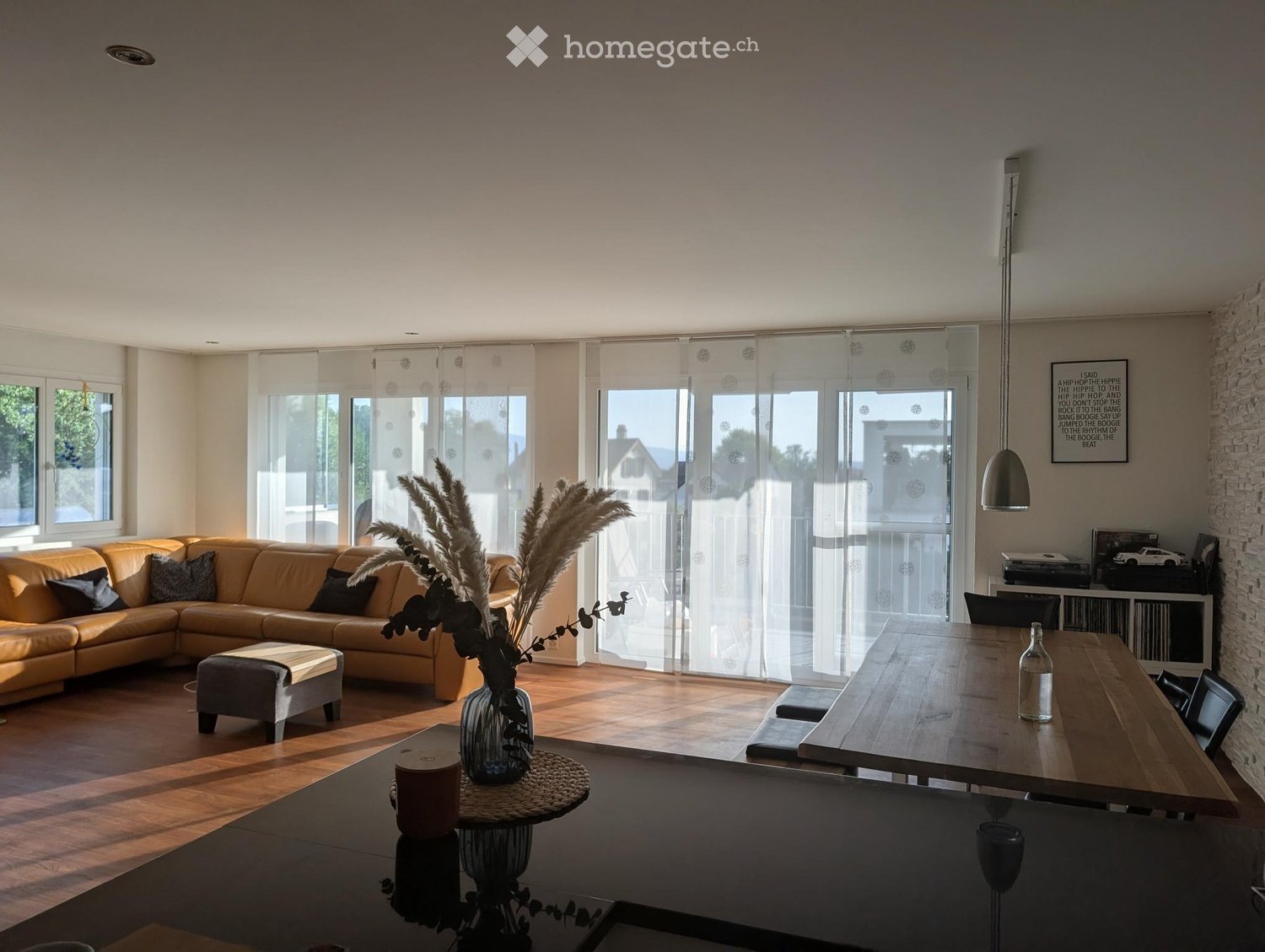 Open-plan living and dining area with a sectional sofa, ottoman, dining table, chairs, and decorative vase with dried plants.