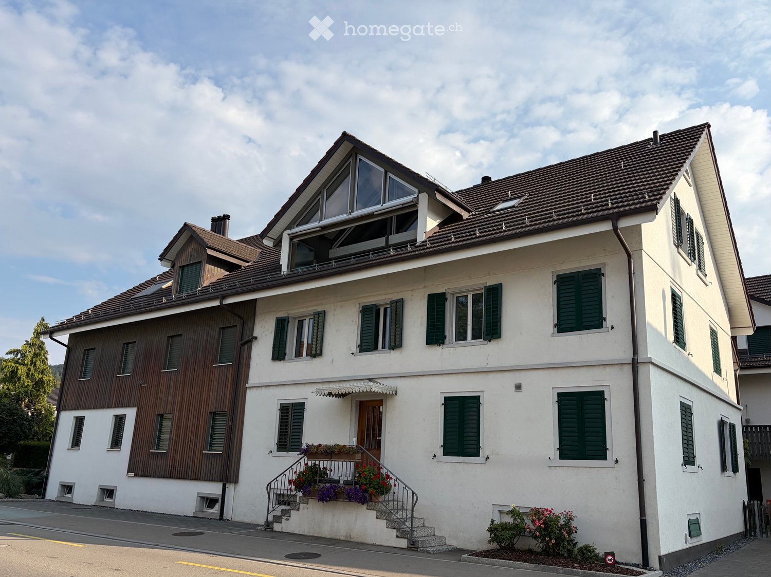 A two-story house with white walls, green shutters, and a brown tiled roof. Features include a front staircase, a small balcony with flower boxes, and a wooden side wall.