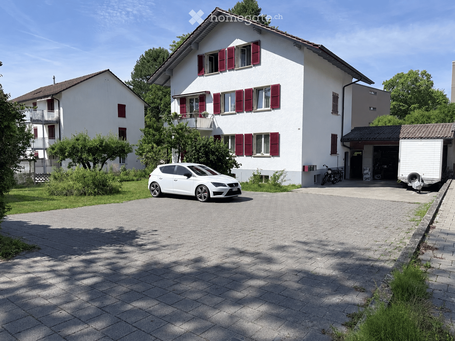 Two-story white house with red shutters, a balcony, and a driveway with a parked car and a motorcycle.