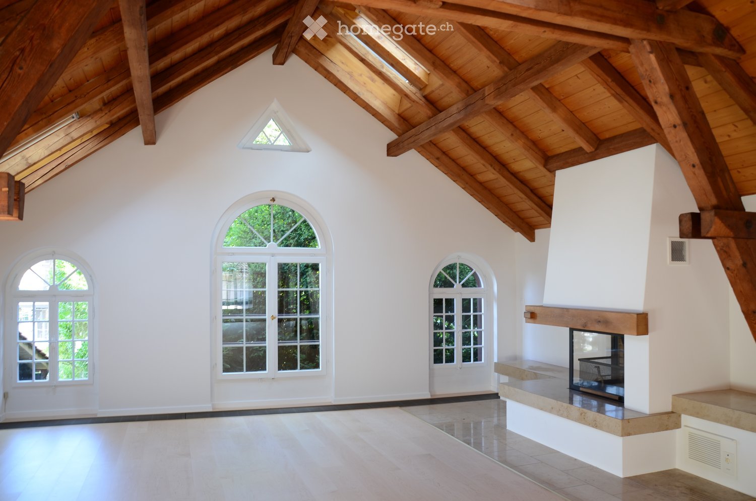 Spacious, high-ceilinged room with exposed wooden beams, white walls, and large arched windows. Features a built-in fireplace with a stone surround and wooden mantel.