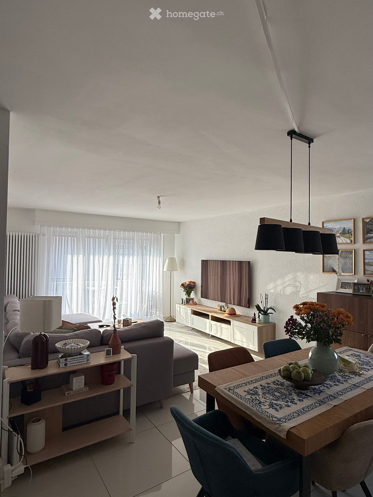 Bright living room with a grey sofa, wooden dining table, chairs, pendant lights, and decorative items including vases and picture frames.