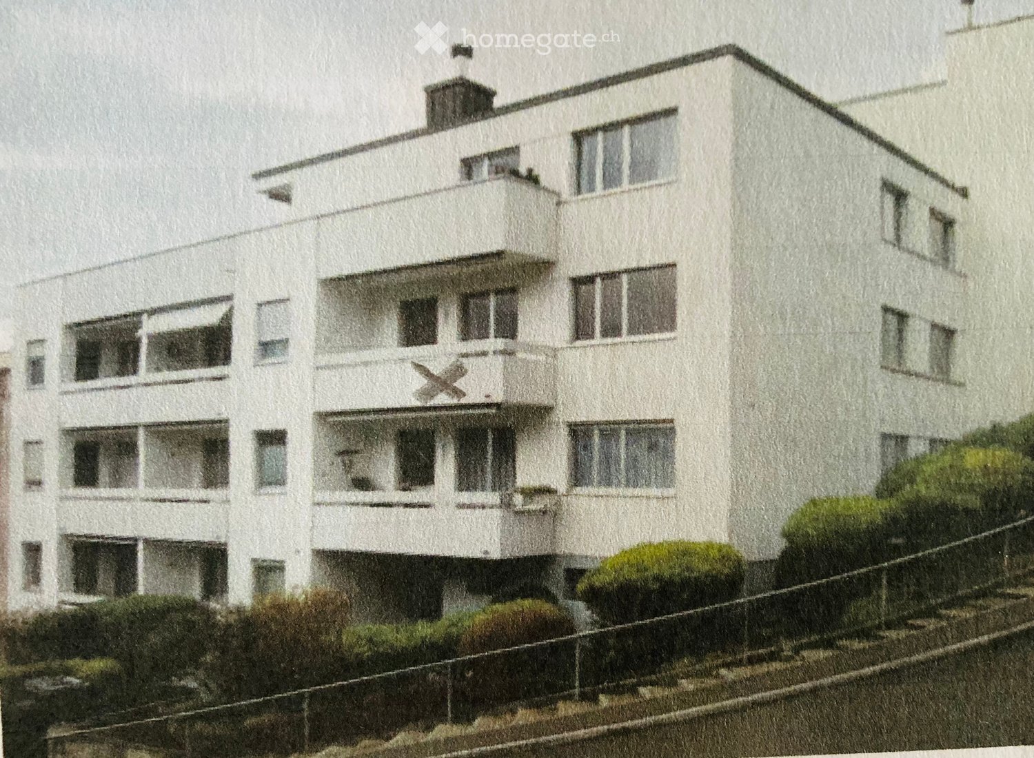 A multi-story white building with numerous windows and balconies, featuring a modern architectural style and a small garden area in front.